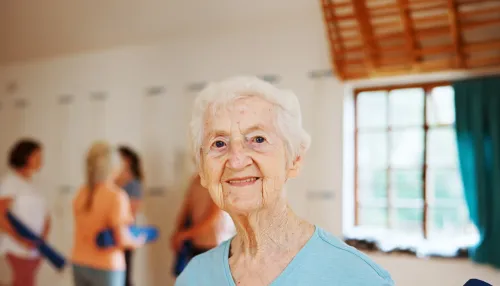Ältere, lächelnde Frau bei der Physiotherapie. Im Hintergrund sind weitere Personen beim Sport zu sehen