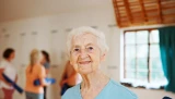 Ältere, lächelnde Frau bei der Physiotherapie. Im Hintergrund sind weitere Personen beim Sport zu sehen