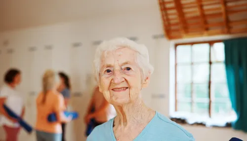 Ältere, lächelnde Frau bei der Physiotherapie. Im Hintergrund sind weitere Personen beim Sport zu sehen