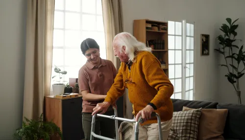 Ein älterer Herr mit Gehilfe in seinem Wohnzimmer wird von eine Pflegekraft beim Aufstehen unterstützt.