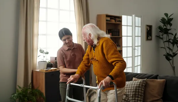 Ein älterer Herr mit Gehilfe in seinem Wohnzimmer wird von eine Pflegekraft beim Aufstehen unterstützt.