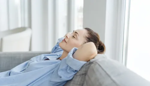 Erschöpfte junge Frau, die sich auf einem Sofa entspannt, mit den Händen hinter dem Kopf und geschlossenen Augen.