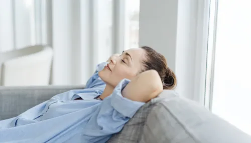 Erschöpfte junge Frau, die sich auf einem Sofa entspannt, mit den Händen hinter dem Kopf und geschlossenen Augen.