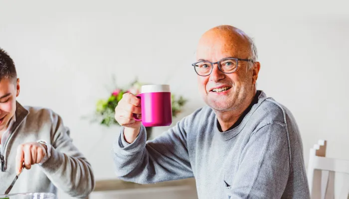Ein älterer Mann mit weißem Haar, der ein blau kariertes Hemd trägt, sitzt auf einem Sofa und schaut nach rechts. Ihm gegenüber steht eine Frau, die ein graues Hemd trägt. Sie reicht ihm einen weißen Teller, auf dem ein in zwei Hälften geschnittener, gelber Apfel liegt.