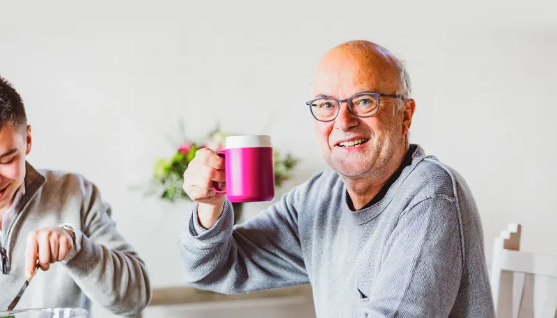 Ein älterer Mann mit weißem Haar, der ein blau kariertes Hemd trägt, sitzt auf einem Sofa und schaut nach rechts. Ihm gegenüber steht eine Frau, die ein graues Hemd trägt. Sie reicht ihm einen weißen Teller, auf dem ein in zwei Hälften geschnittener, gelber Apfel liegt.