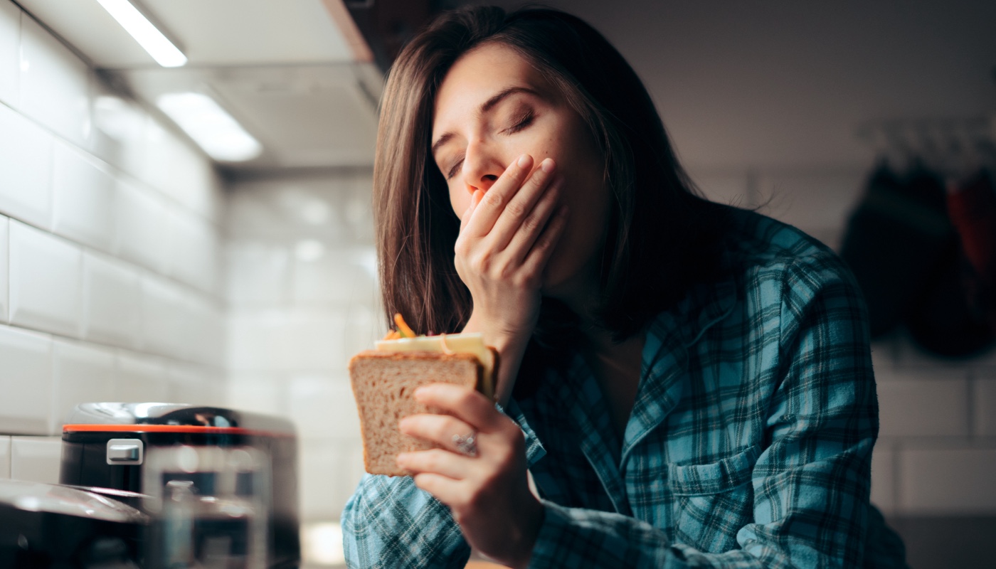 Gähnende junge Frau in der Küche, die ein Käsetoast in der Hand hält